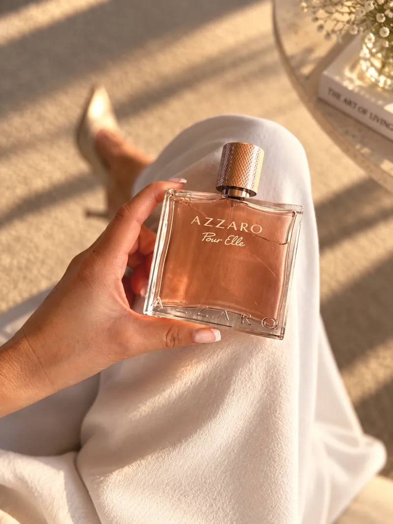 Le flacon Azzaro Pour Elle tenu à la main au-dessus d’une robe blanche, dans une lumière rasante de fin de journée